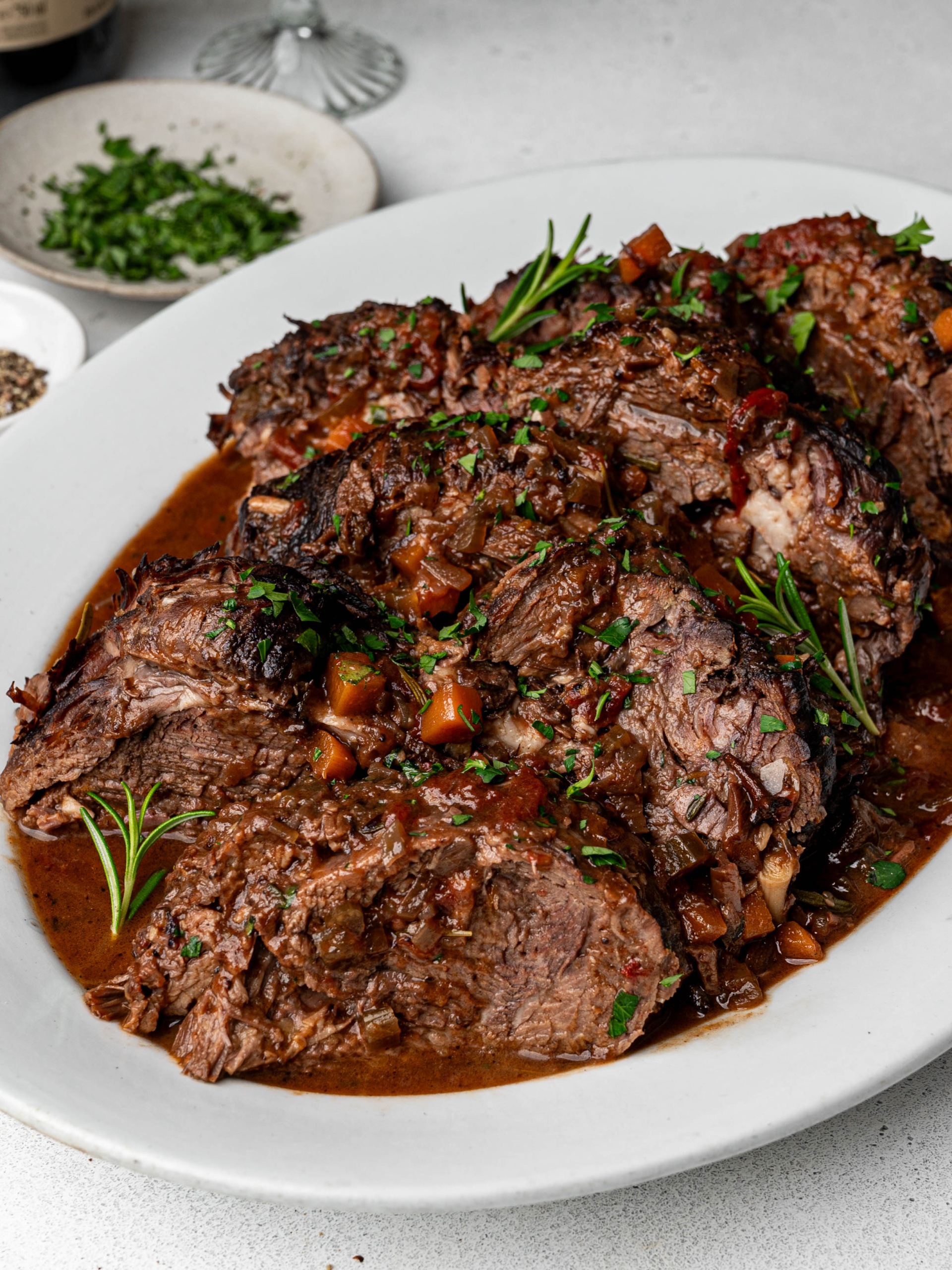 Big chunks of braised Stracotto di Manzo served on a white platter garnished with chopped parsley rosemary sprigs.