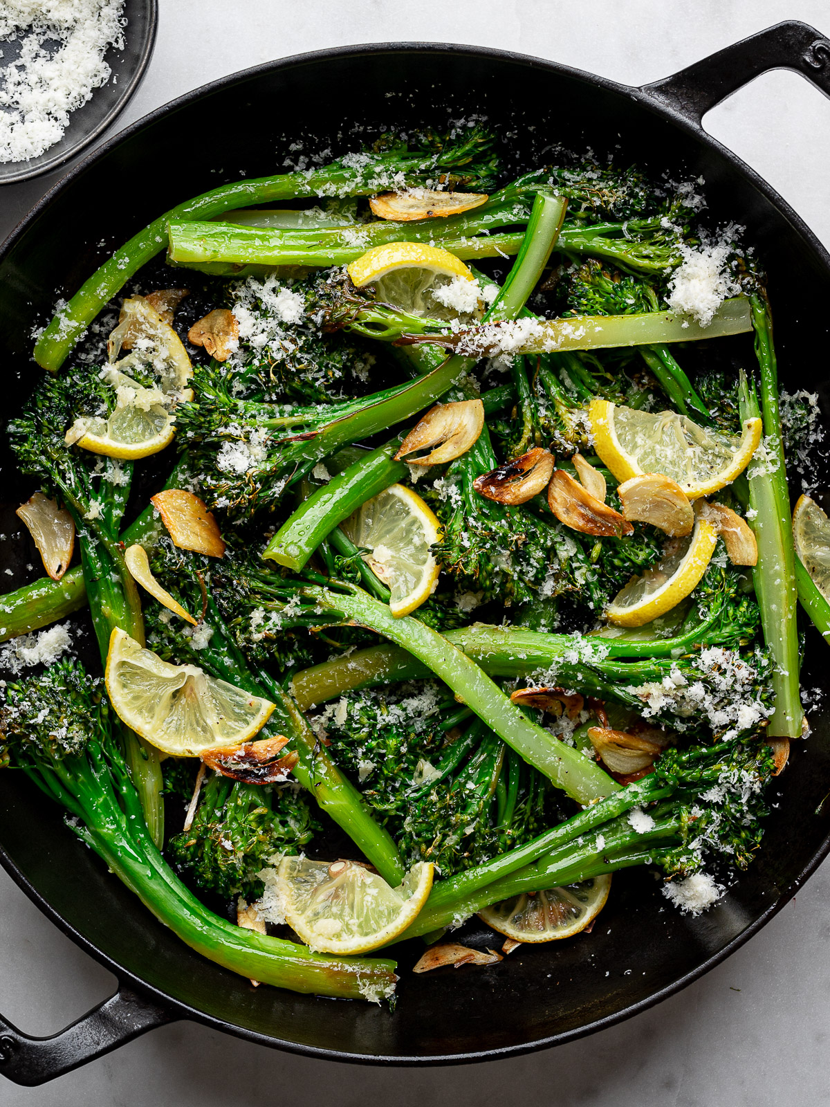 Broiled broccolini stalks in black skillet with lemon slices, parmesan cheese and garlic chips. 