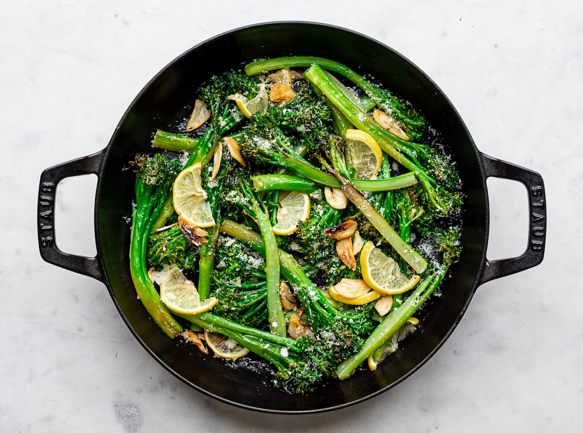 Broiled broccolini stalks in black skillet with lemon slices, parmesan cheese and garlic chips.