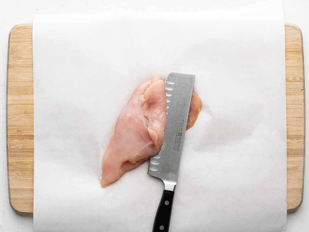 One chicken breast getting sliced in half with a sharp knife to make cutlets. 