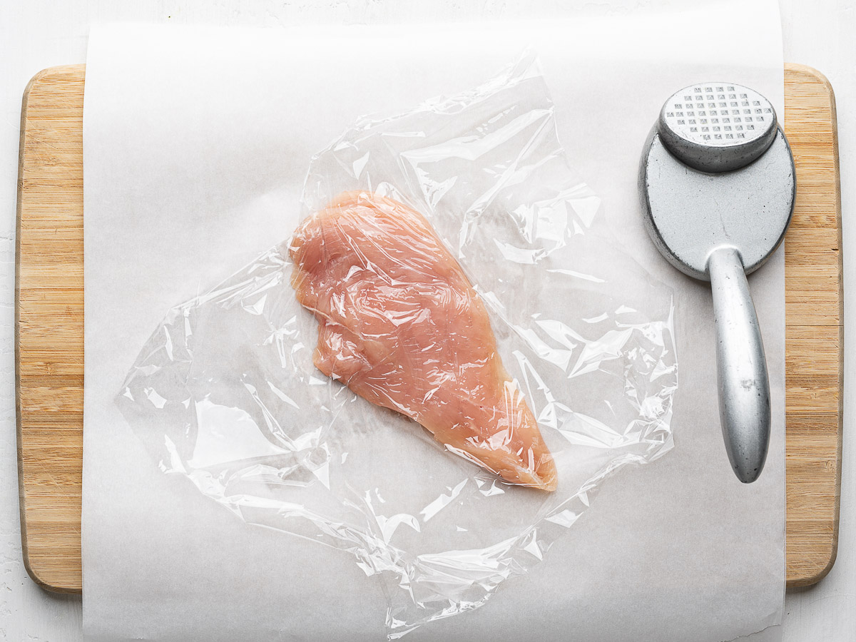 Pounding one chicken breast on a cutting board to make it thinner. 