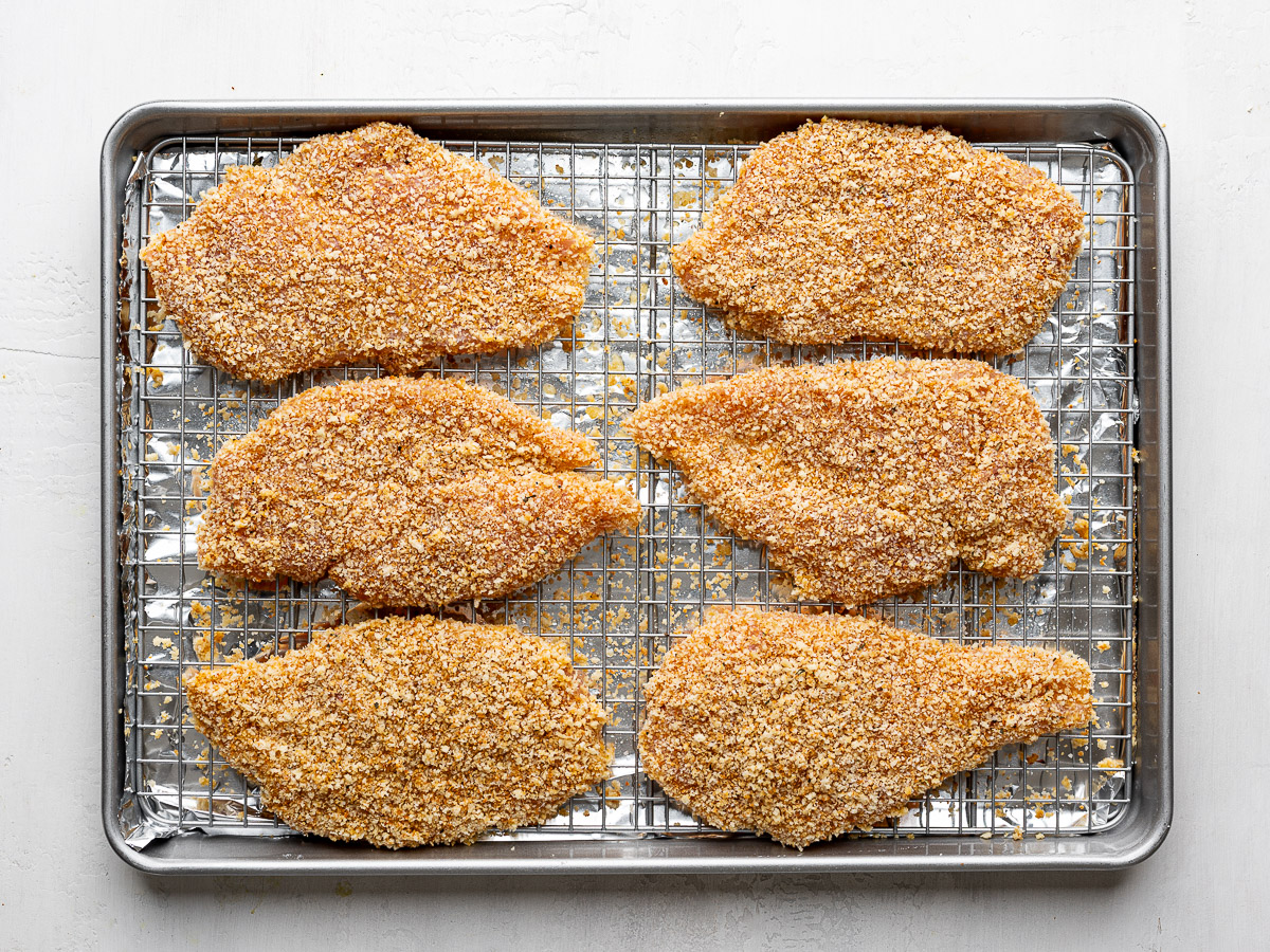 Six un-baked chicken cutlets coated in panko on a wire rack set over a sheet pan.