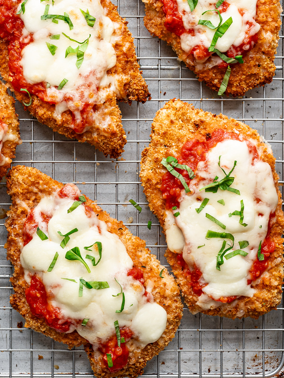 Close-up of baked chicken parmesan cutlets on a wire rack with marinara sauce, melty cheese and garnished with fresh basil.