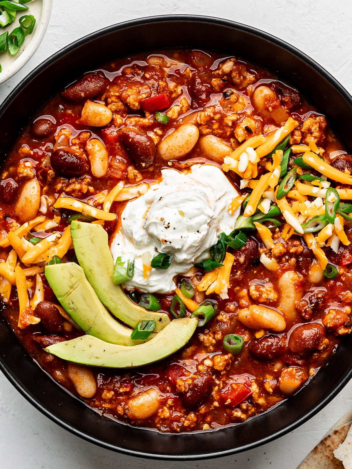 Close up of harissa turkey chili served in a black bowl and topped with avocado slices, sour cream and grated cheddar.