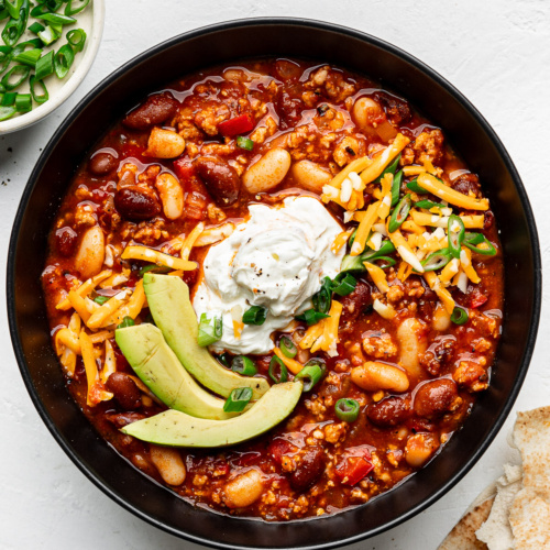 Harissa turkey chili served in a black bowl and topped with avocado slices, sour cream and grated cheddar.