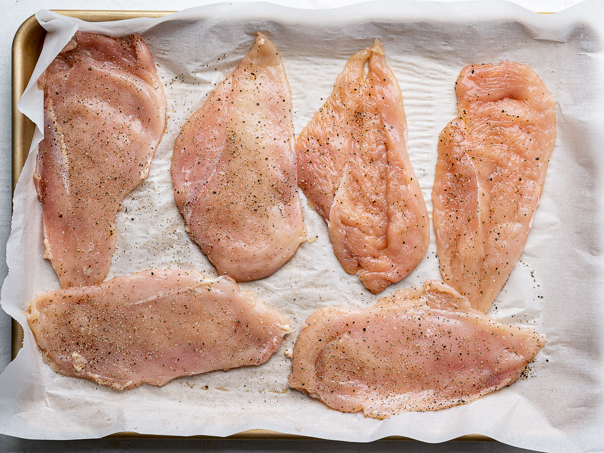 Six un-baked chicken cutlets on a baking sheet seasoned with salt and pepper. 