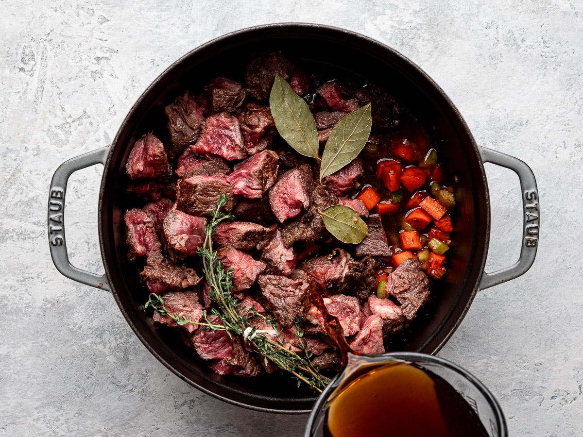 Pouring broth over vegetable mixture and chopped beef pieces in Dutch Oven.  
