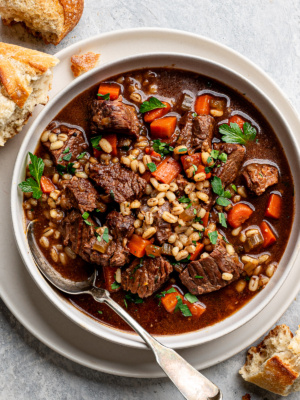 Beef barley soup served in white shallow bowl with crusty bread on the side.