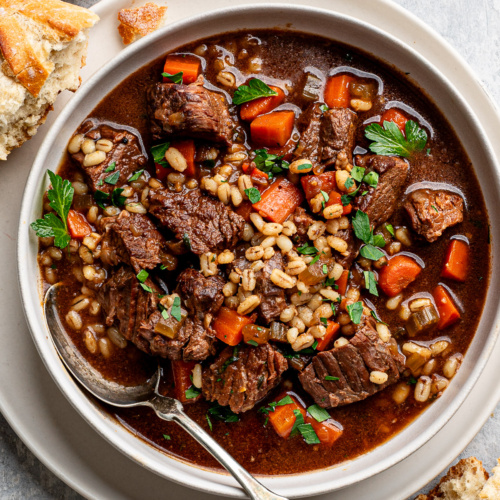 Beef barley soup served in white shallow bowl with crusty bread on the side.