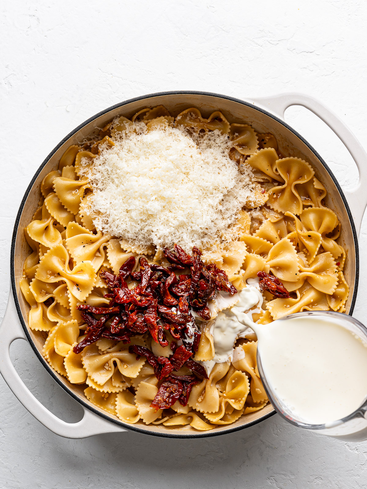 Adding sun-dried tomatoes, parmesan, and heavy cream to cooked pasta in skillet.