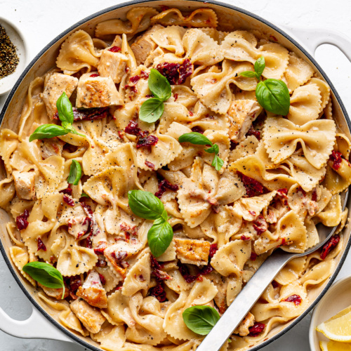 The finished dish of marry me chicken pasta in a large white skillet topped with fresh basil and lemon wedges on the side.