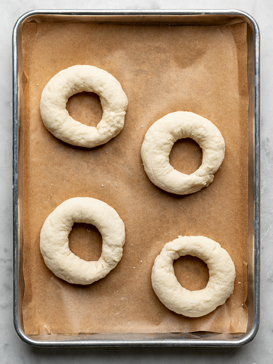 4 shaped raw bagels on baking sheet. 