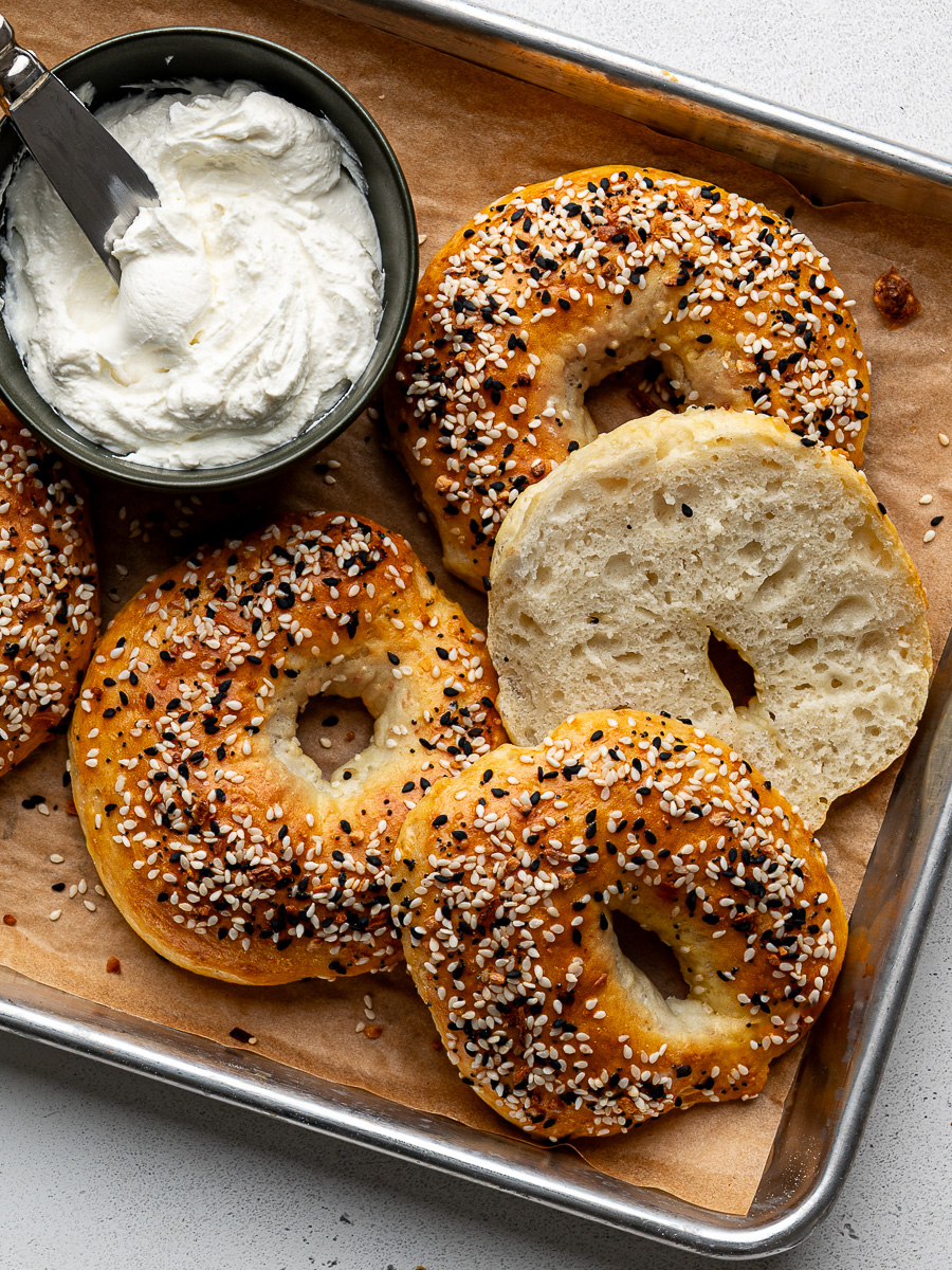 A sliced bagel with other bagels on baking sheet with cream cheese on the side. 