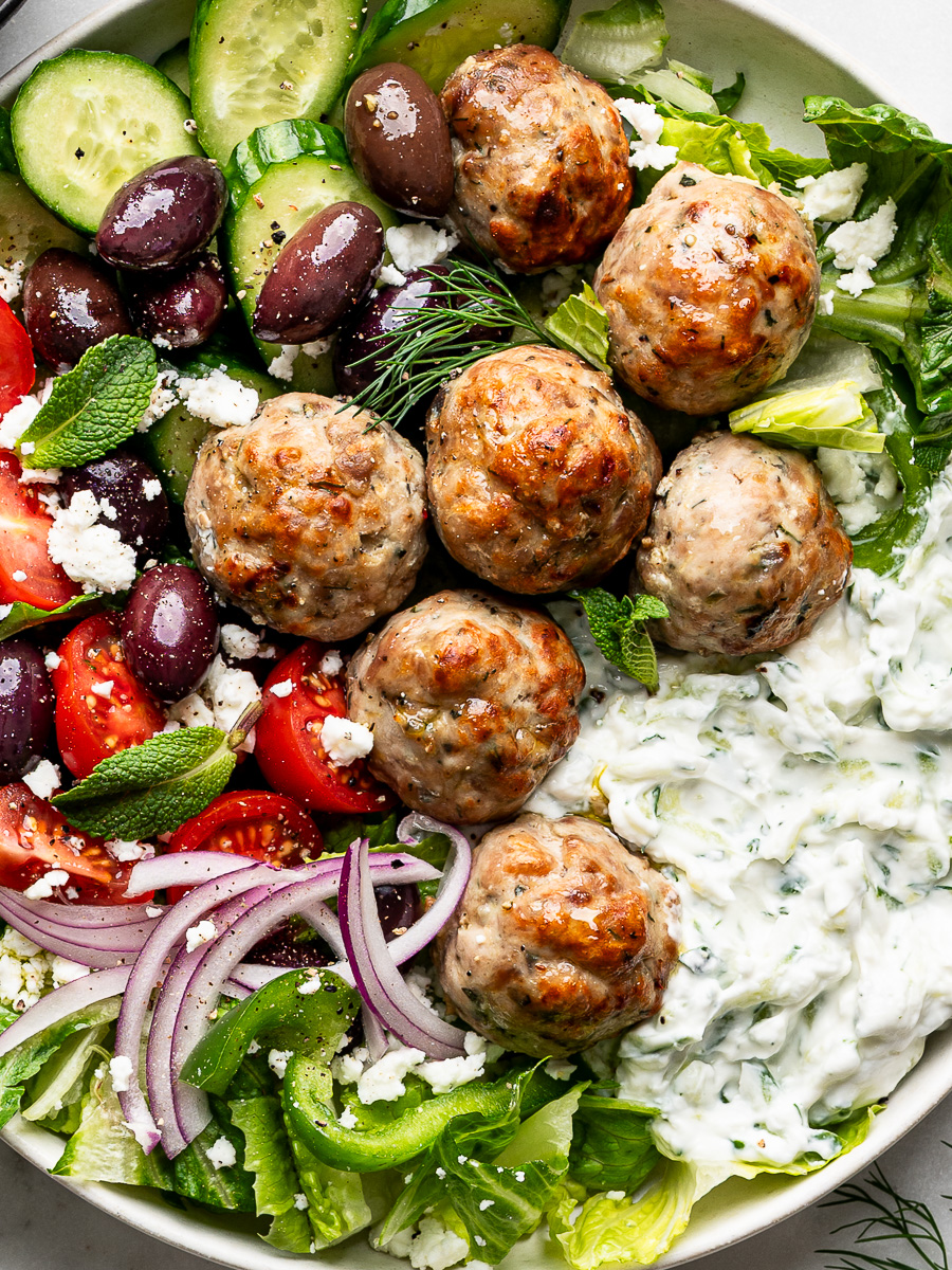 Close-up of Greek Chicken meatballs served in a white bowl with tzatziki sauce and a Greek salad.