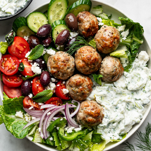 Greek Chicken meatballs served in a white bowl with tzatziki sauce and a Greek salad.