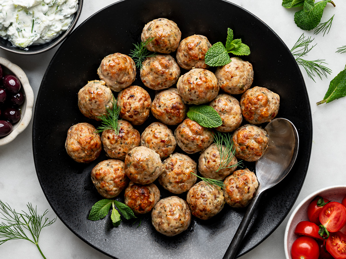 Baked Greek chicken meatballs on a black serving bowl with bowls of tzatziki sauce, Kalamata olives, and cherry tomatoes on the side. 
