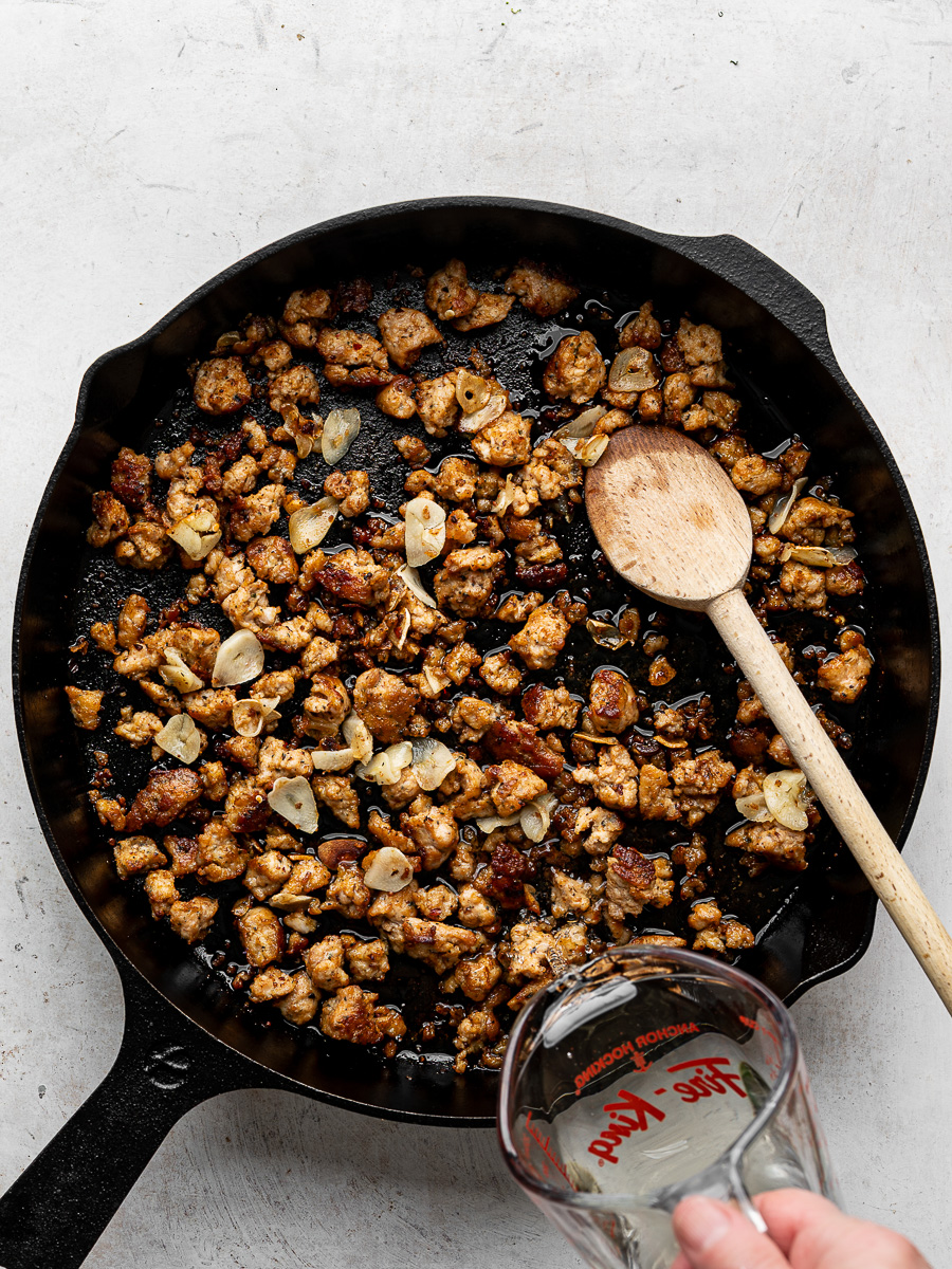 Golden brown crumbled Italian sausage in a black skillet with thinly sliced garlic. 