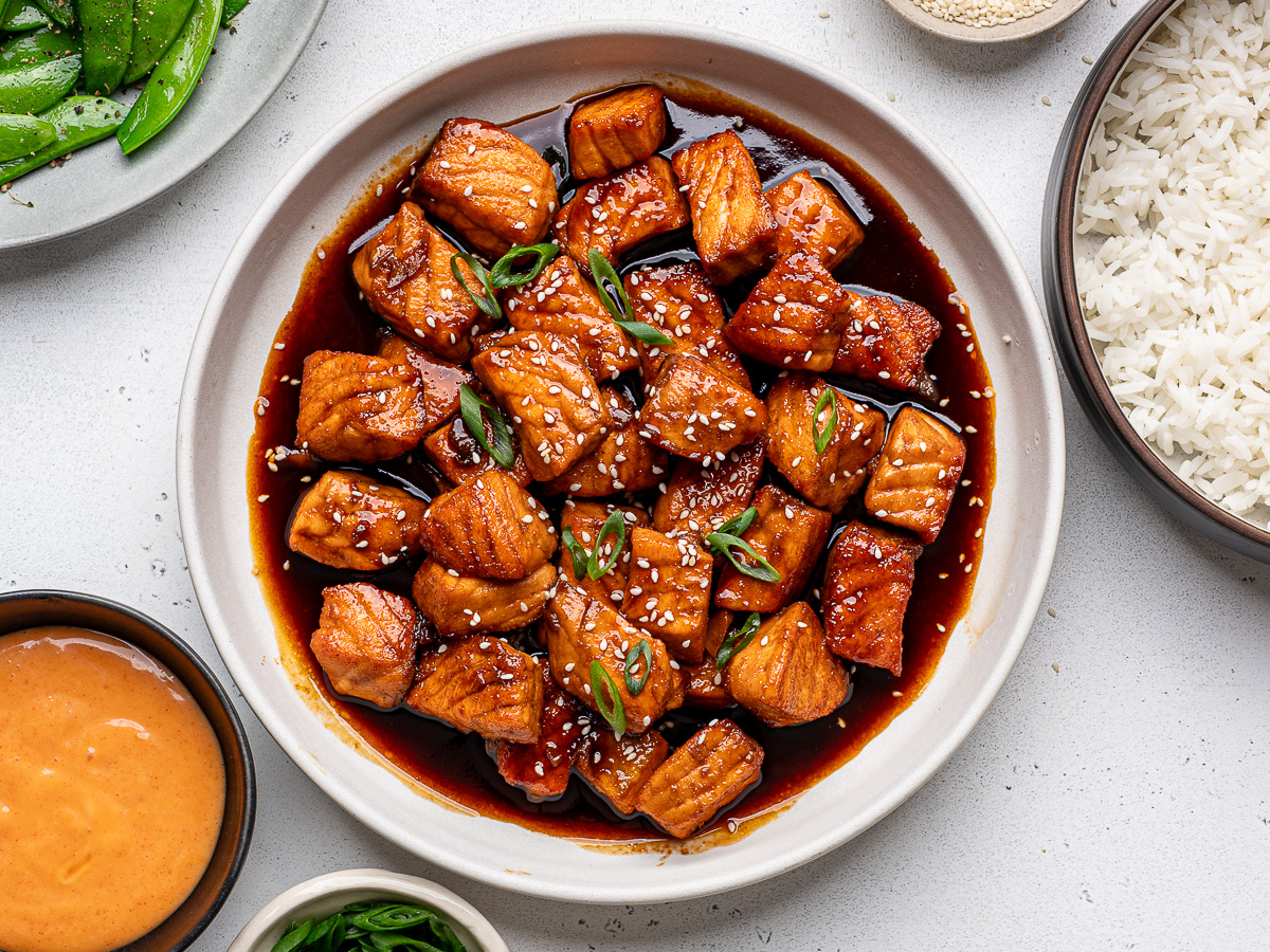 Cooked salmon bites in shallow white bowl garnished with sesame seeds and green onions and bowls of rice, sugar snap peas and sauce on the side. 