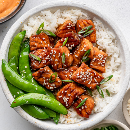 Honey glazed salmon bites served in a white bowl over rice with steamed sugar snap peas and a drizzling sauce on the side.