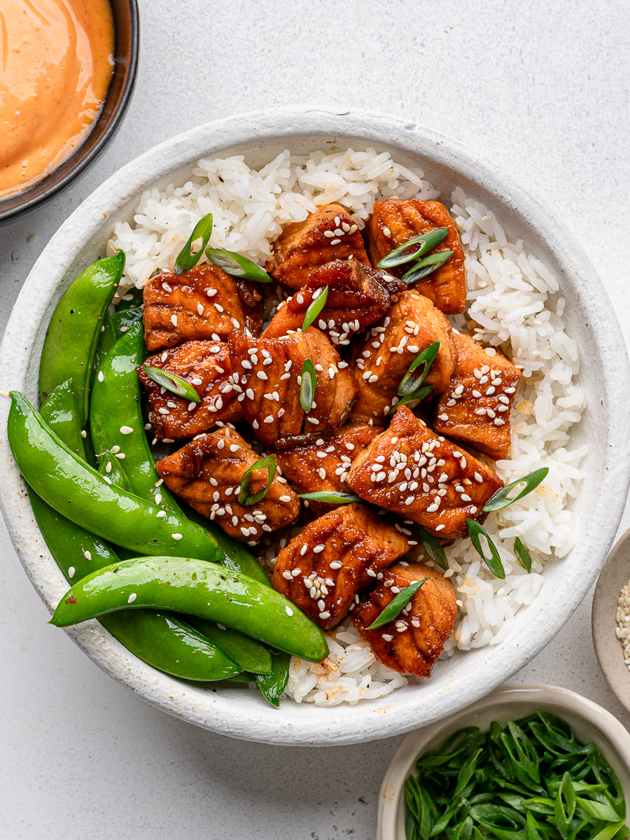 Honey glazed salmon bites served in a white bowl over rice with steamed sugar snap peas and a drizzling sauce on the side.