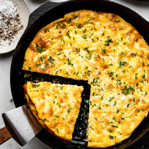 Golden brown ham and cheese frittata in a cast iron skillet with spatula removing a piece ready to serve.