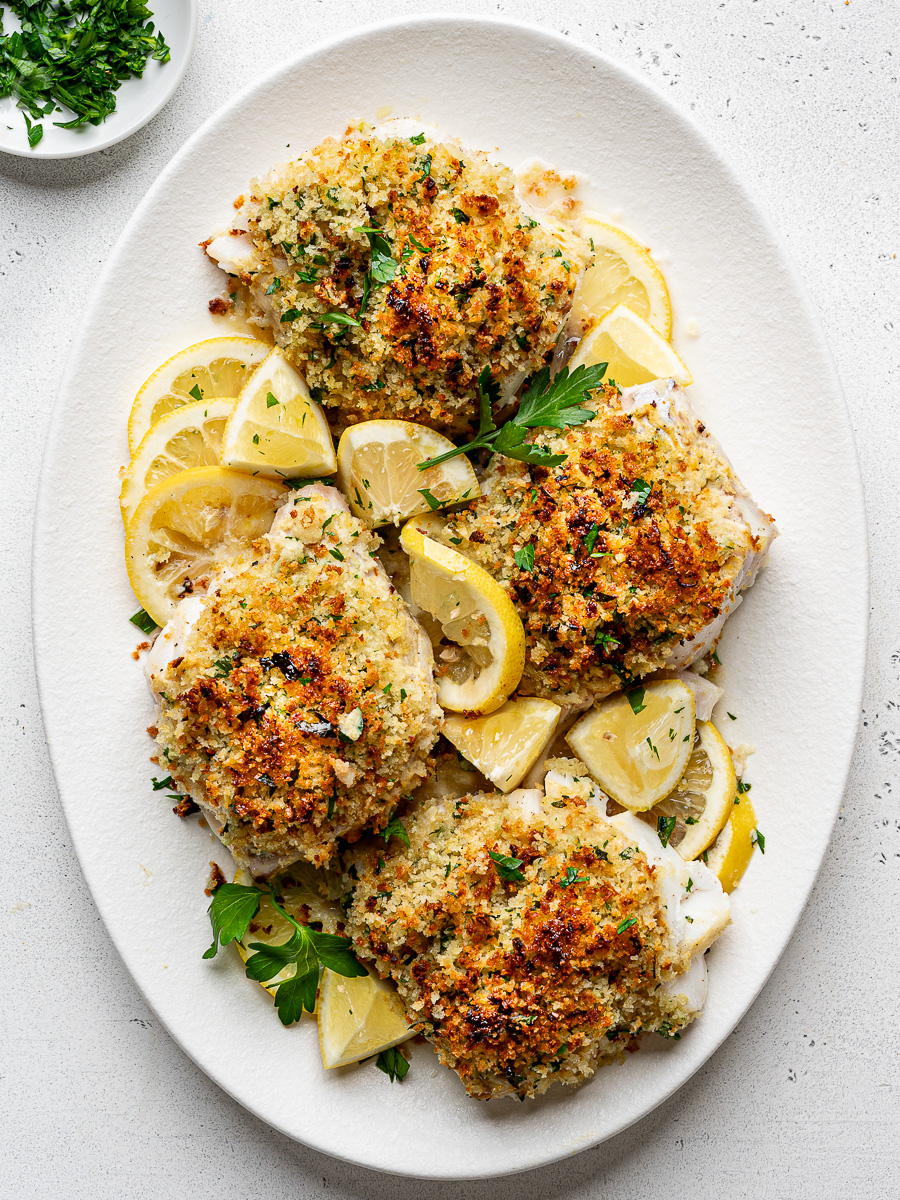 Four golden brown panko crusted cod fillets served with lemon wedges and fresh herbs on a white platter.