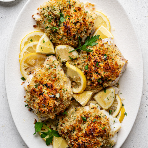 Four golden brown panko crusted cod fillets served with lemon wedges and fresh herbs on a white platter.