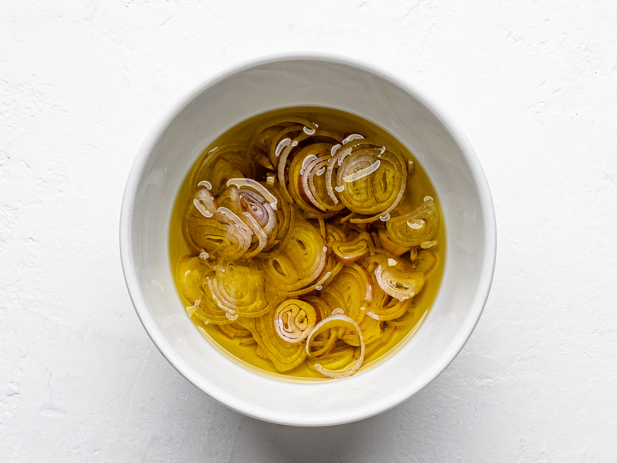 Sliced raw shallots in small white bowl covered with olive oil. 