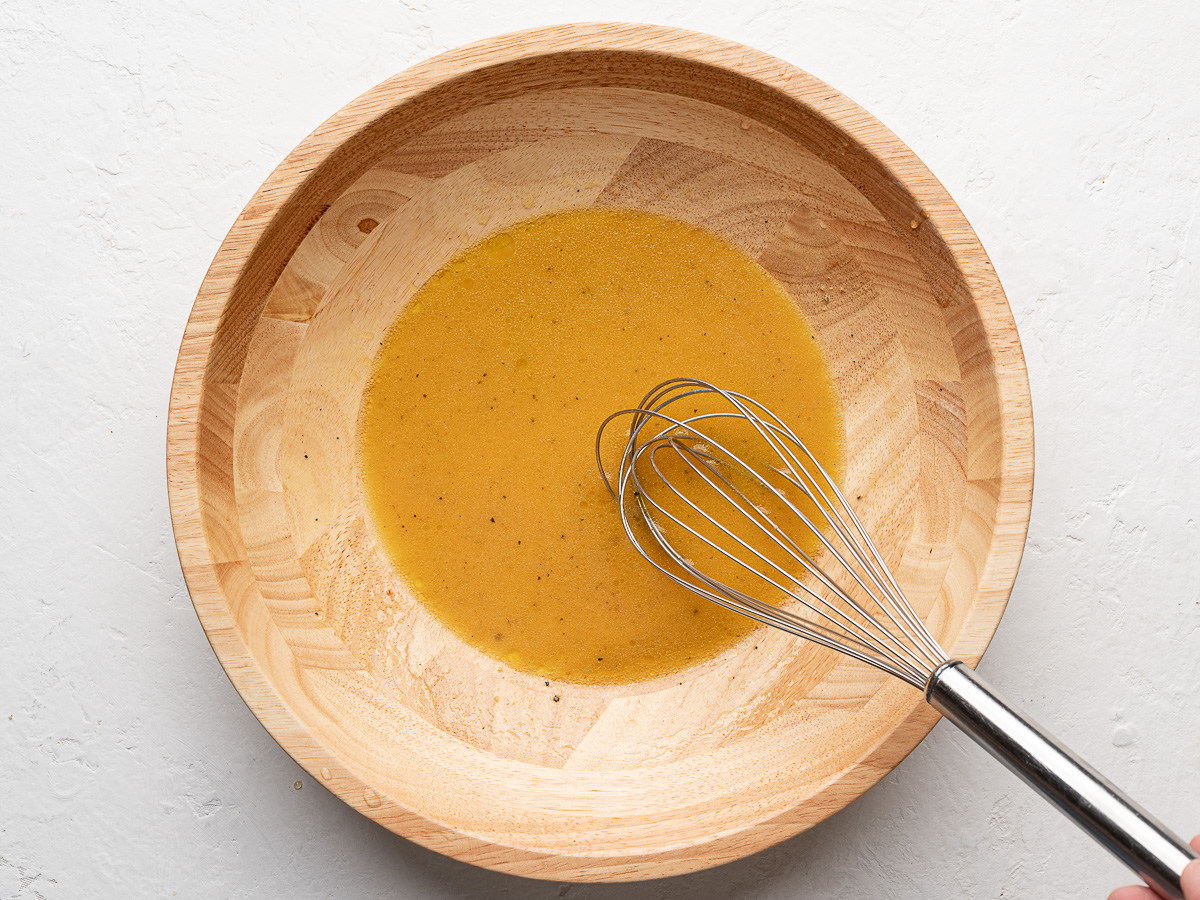 Salad dressing ingredients mixed in wooden bowl with whisk.
