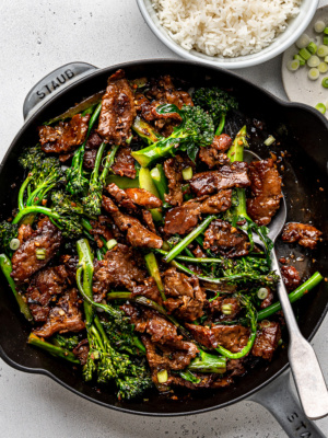 Close-up of cooked beef and Broccolini stir-fry in large cast-iron skillet with bowl of rice on the side.