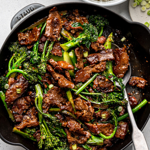 Close-up of cooked beef and Broccolini stir-fry in large cast-iron skillet with bowl of rice on the side.