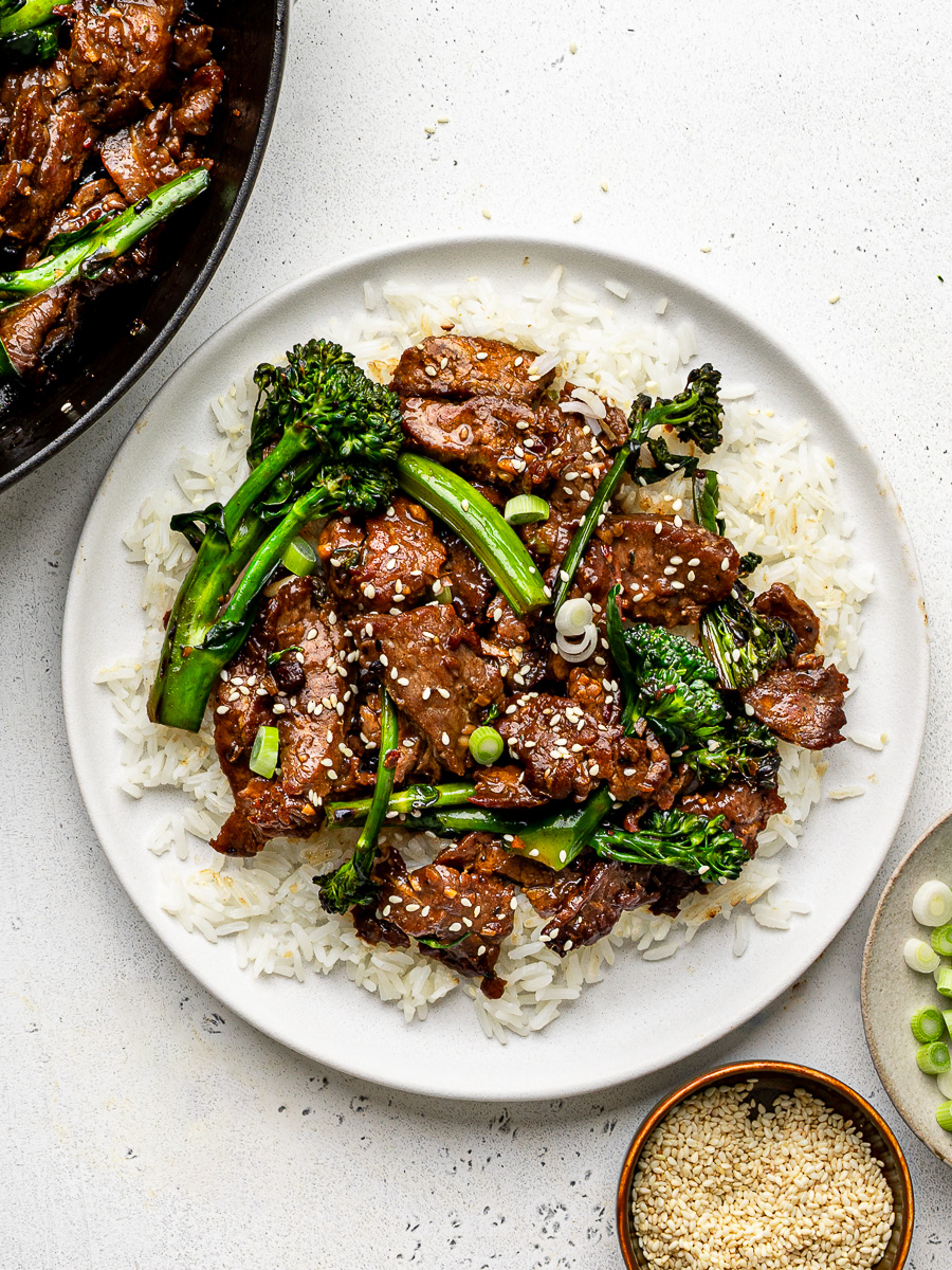 Stir-fry served over white rice on a white plate with skillet beside it. 