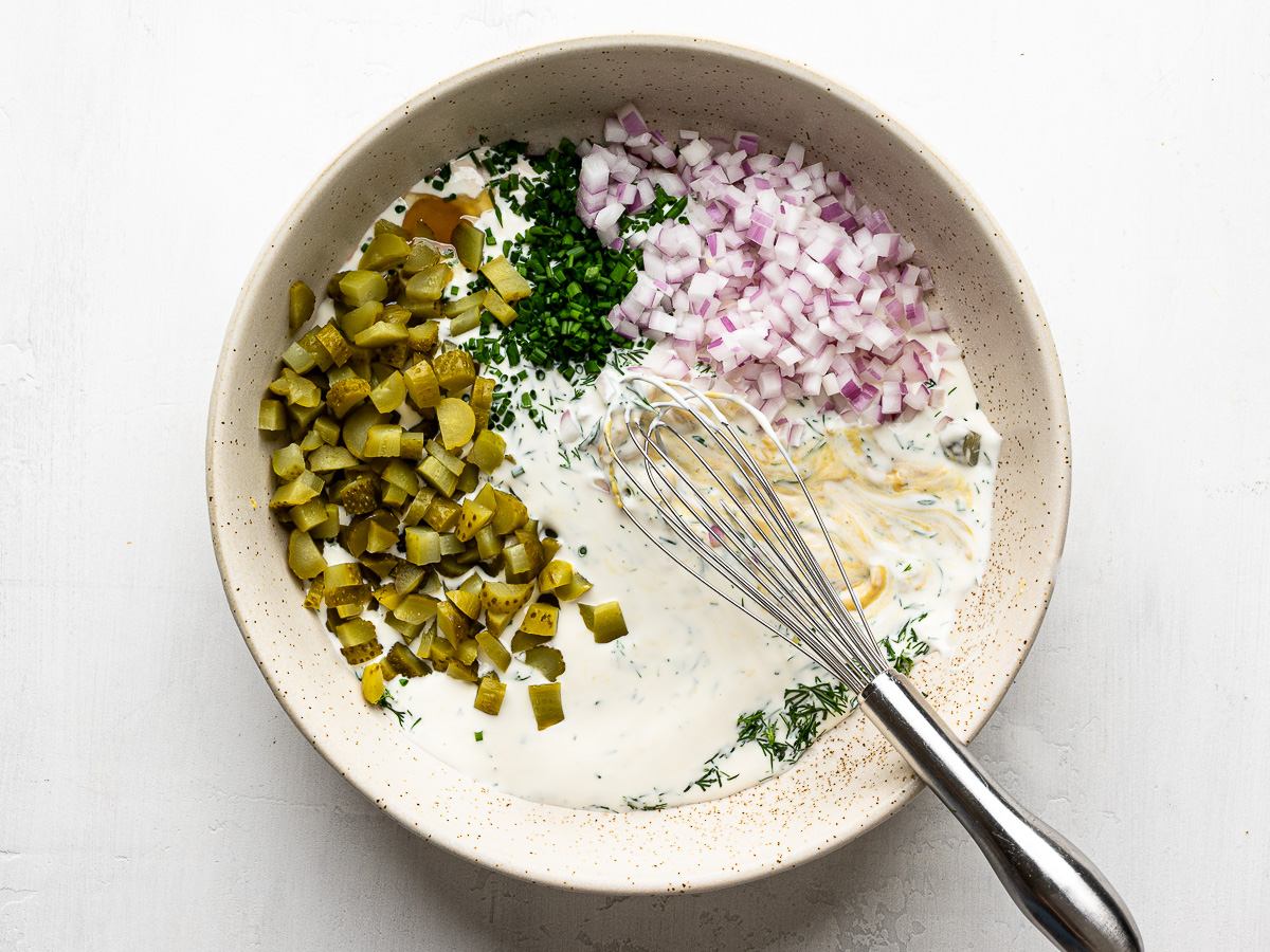 Mixing dressing for smashed potatoes made with Greek yogurt, mayo, lemon juice, dill, red onions, and chopped pickles in a salad bowl.