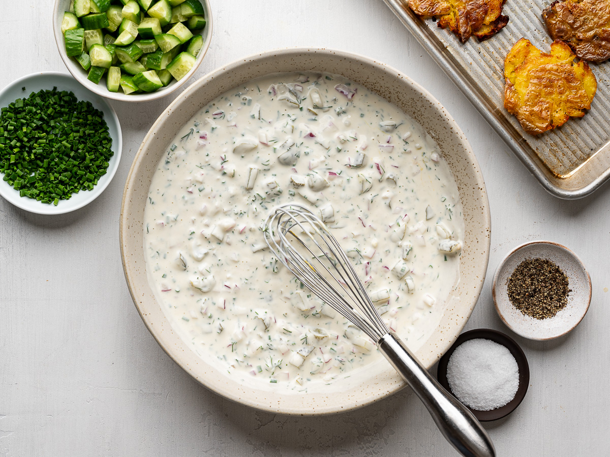 Creamy dressing made with Greek yogurt, mayo, lemon juice, dill, and chopped pickles in a mixing bowl and showing baking sheet with roasted smashed potatoes.