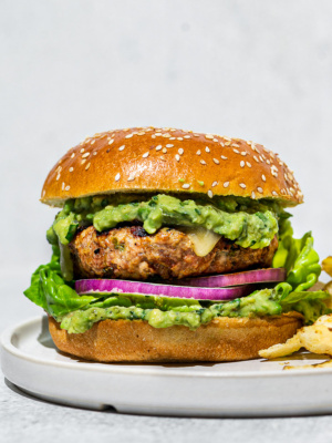 One poblano turkey burger filled with lettuce, red onion slices, and avocado sauce on a white plate with chips on the side.