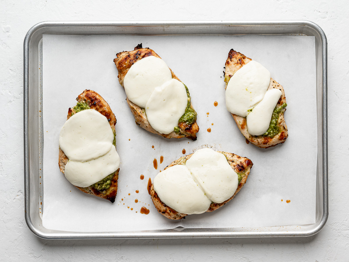 Grilled chicken breasts topped with basil pesto and melting fresh mozzarella cheese on a baking sheet.