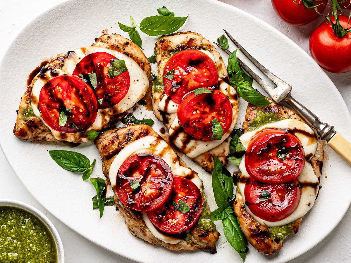 Grilled Caprese chicken on a platter topped with tomato slices, fresh basil, and a drizzle of balsamic glaze.