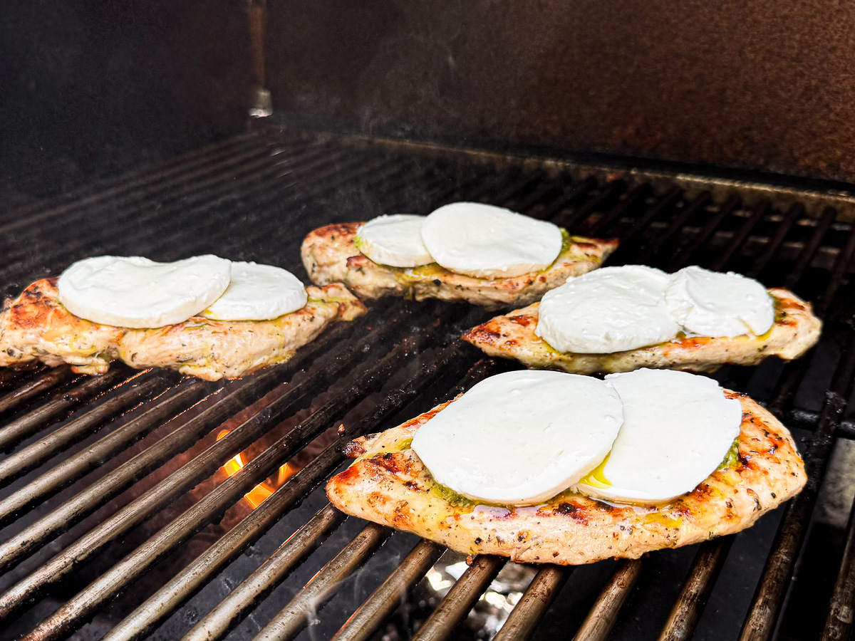 Grilled chicken breasts on the grill topped with basil pesto and melting fresh mozzarella cheese.