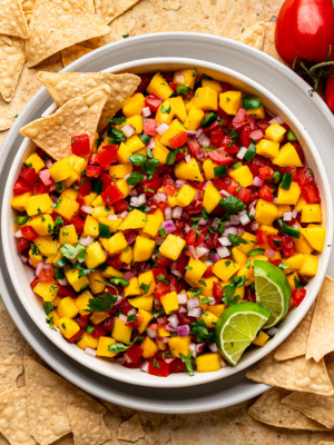 A bowl of mango pico de gallo salsa served in a white bowl with tortilla chips and limes.