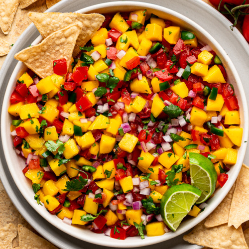 A bowl of mango pico de gallo salsa served in a white bowl with tortilla chips and limes.