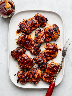 Grilled BBQ boneless chicken thighs on a large white platter with extra BBQ sauce on the side.