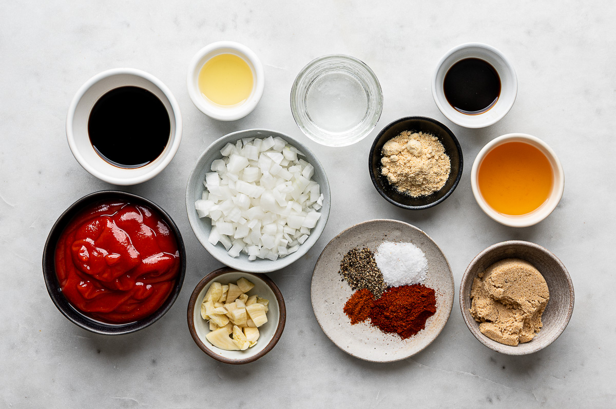 Recipe ingredients for homemade BBQ sauce prepared in bowls which include ketchup, chopped onion, chopped garlic, and balsamic vinegar.