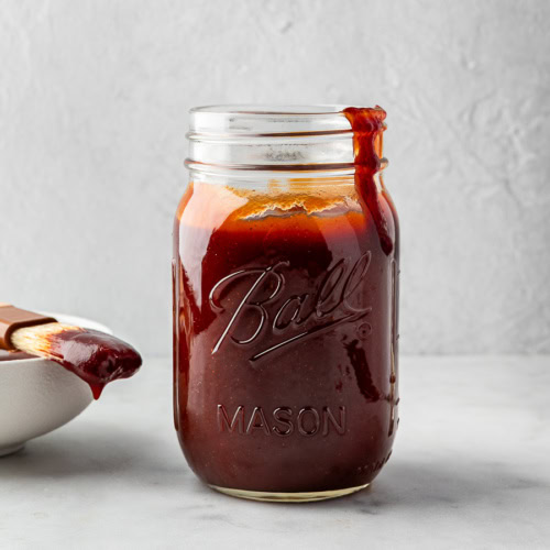 Homemade BBQ sauce in mason jar with small white bowl and pastry brush beside it.