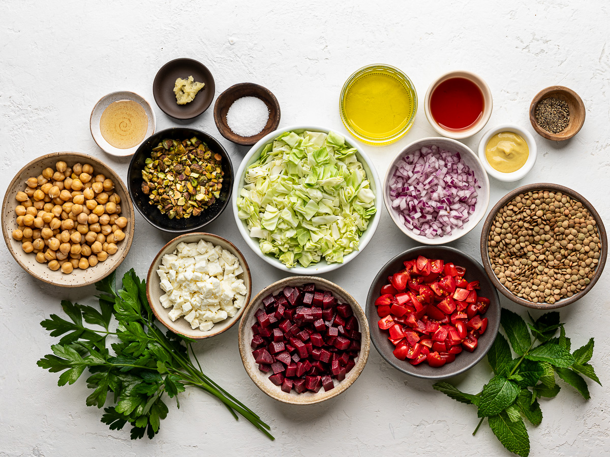 Ingredients for lentil chopped salad including green lentils, chopped cabbage, cherry tomatoes, red onion, fresh herbs, olive oil, lemon, feta, and spices—everything you need for a healthy, vegetarian salad recipe.