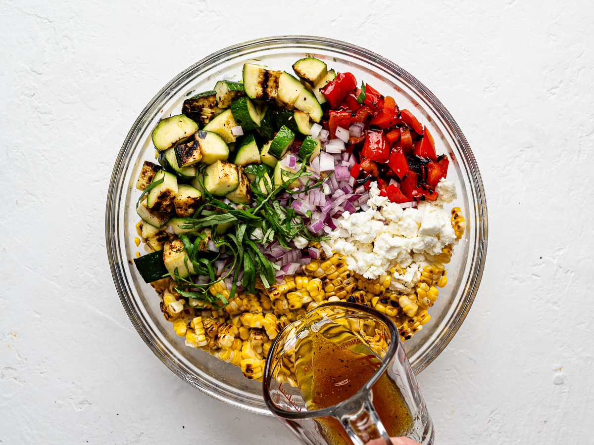 Pouring vinaigrette over salad ingredients in glass mixing bowl.