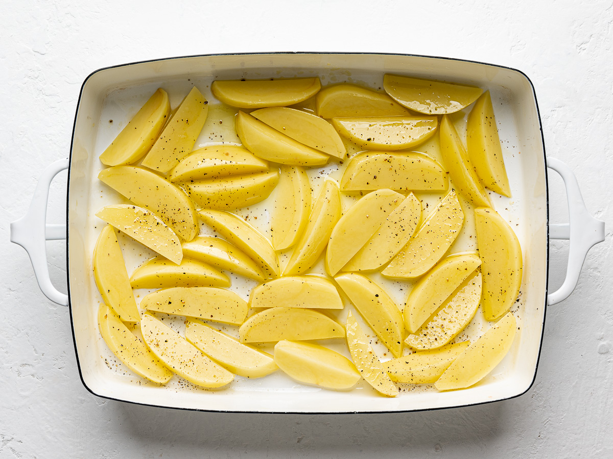 Seasoned potato wedges spread in a single layer in a white baking dish, ready for par-roasting.