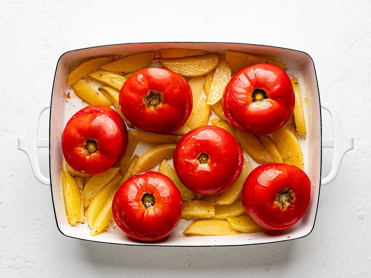 Tomatoes filled with rice mixture sitting atop potato wedges with tops on before baking.