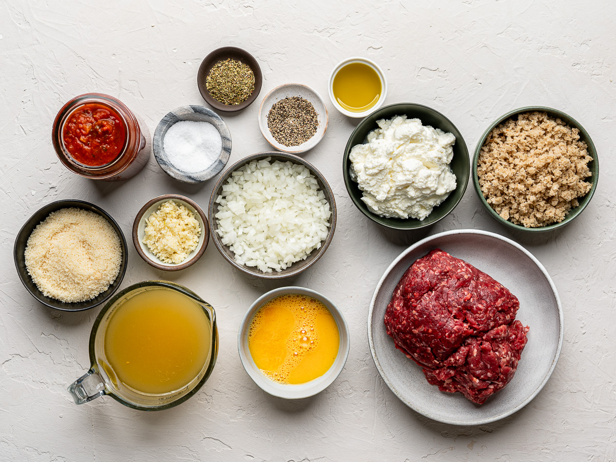 Ingredients for ricotta meatballs arranged on a kitchen counter: ground beef, ricotta, onion garlic, Parmesan and breadcrumbs.