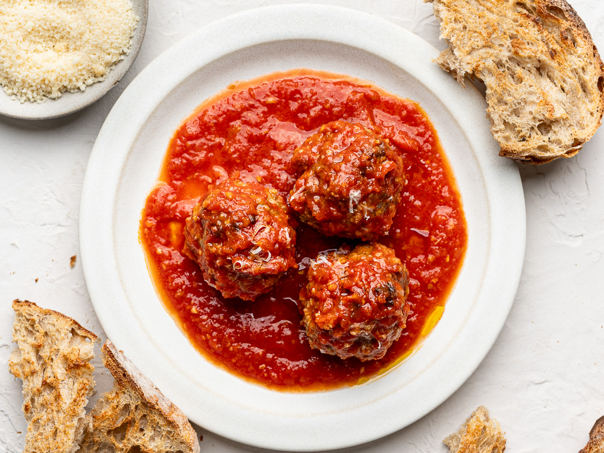 Closeup of three meatballs served on a white plate with crusty bread on the side.