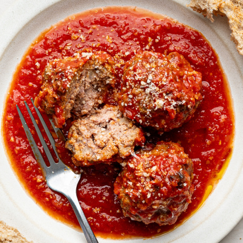 Closeup of three meatballs served on a white plate with crusty bread on the side.